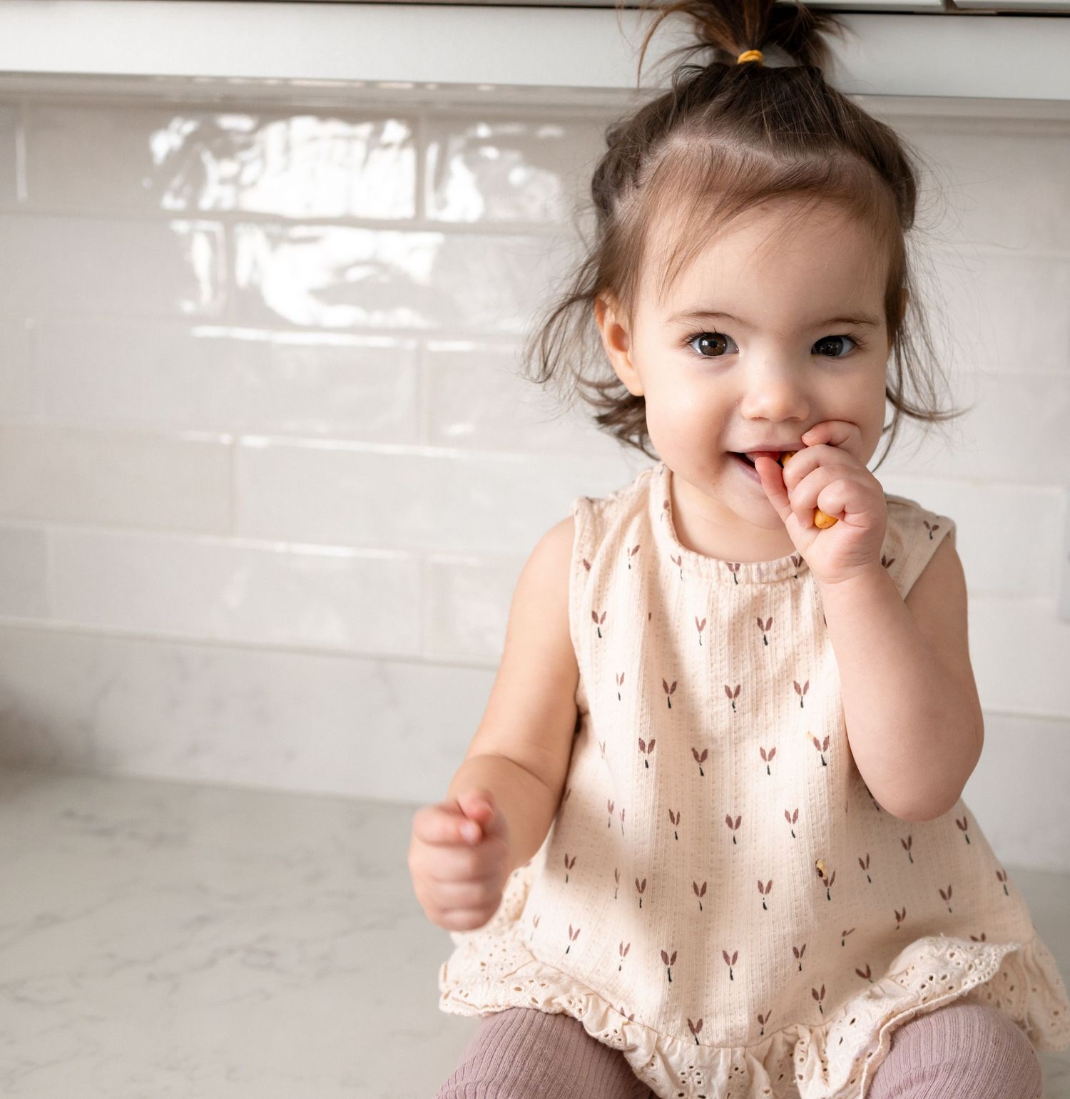 Adorable toddler girl in a cream floral romper sitting on a kitchen counter, smiling and eating a snack. Photography in Buckinghamshire.