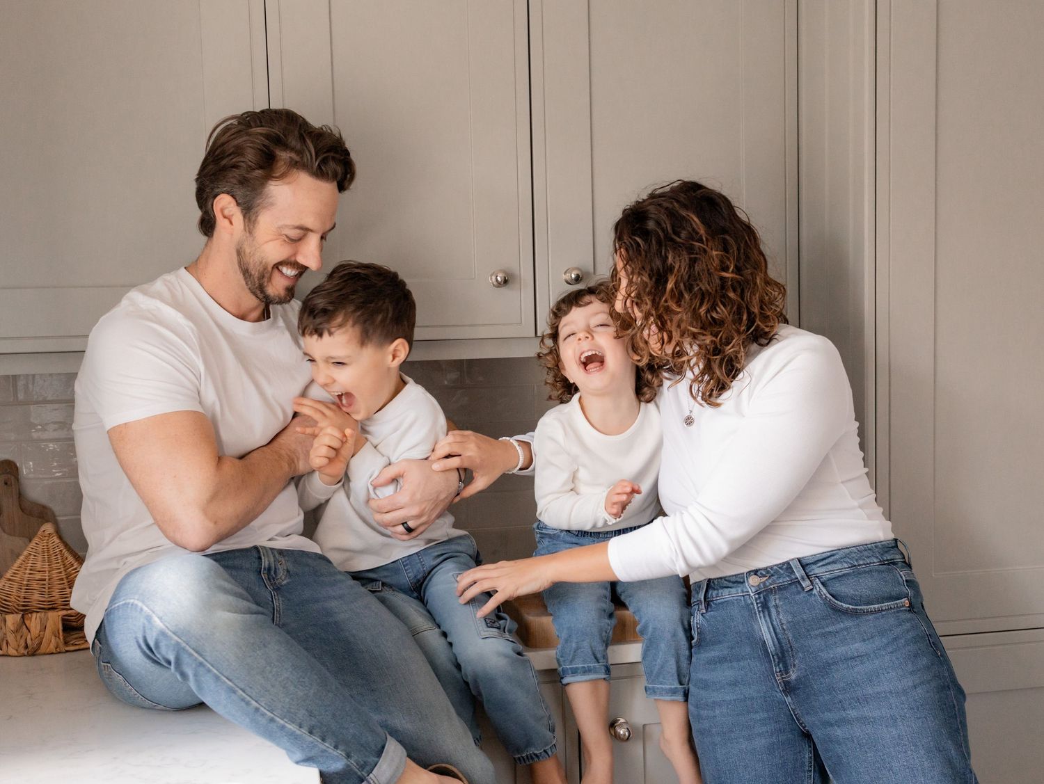 Family members wearing jeans and white shirts playfully interact while sitting on a grey couch together. Photography in Buckinghamshire.