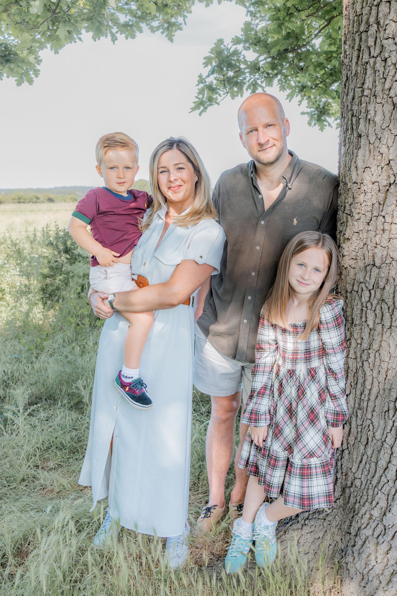 Family of four posing by a tree outdoors, parents with young boy and girl in summer clothes.