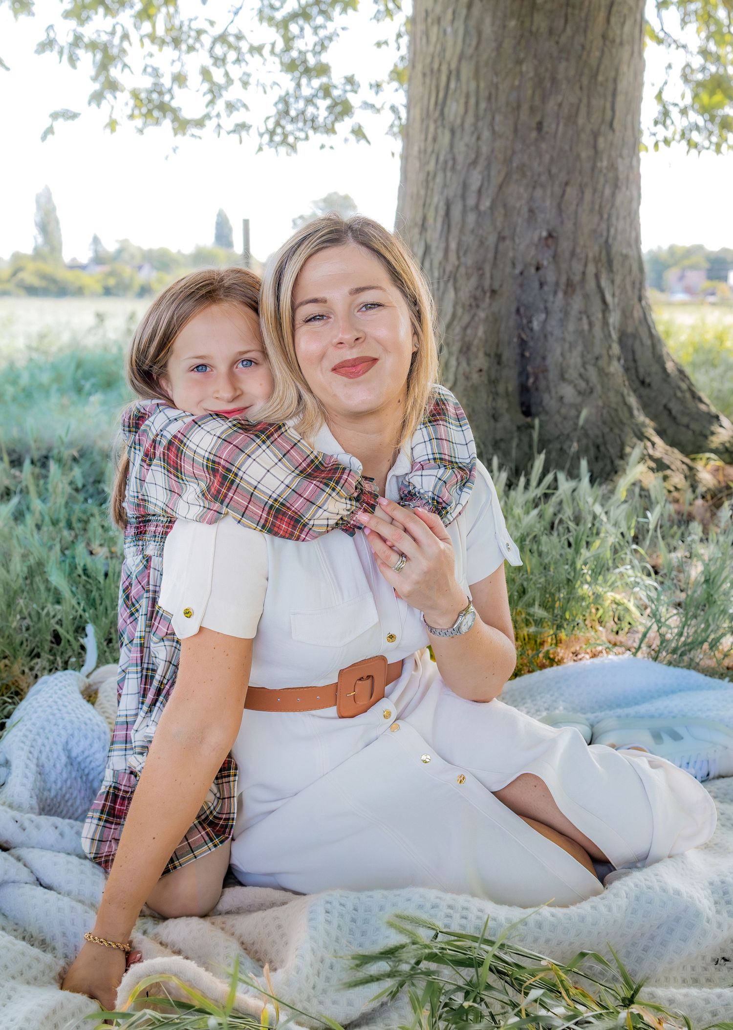 A woman and young girl share a warm embrace on a blanket under a tree during an outdoor picnic.