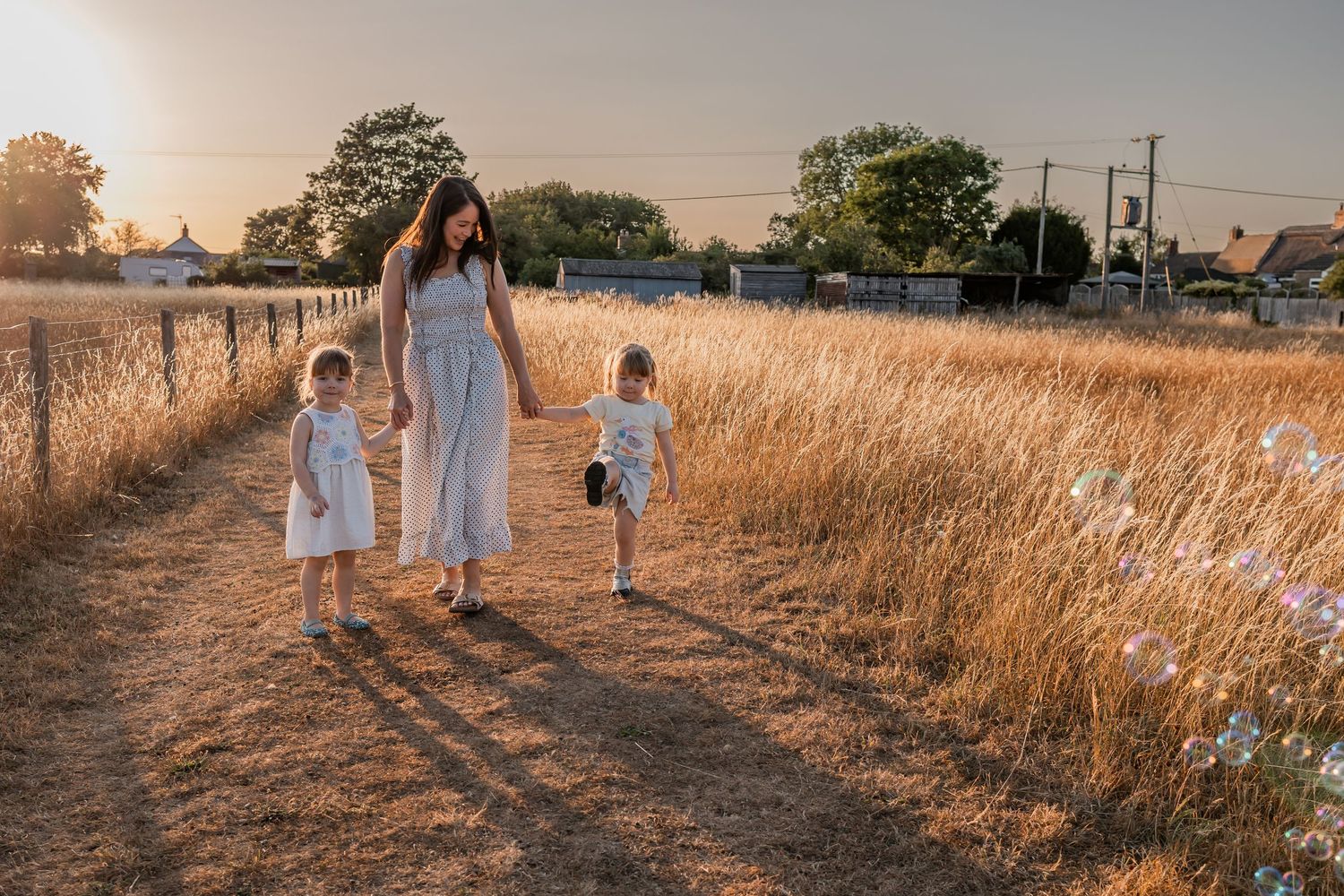 A mother and two children walk along a golden field path at sunset, all wearing white dresses in a serene rural setting.