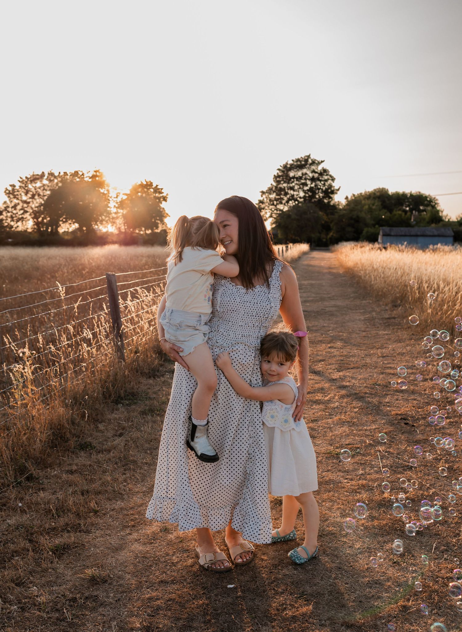 A mother holds a toddler while another child hugs her on a golden sunset path in a field.