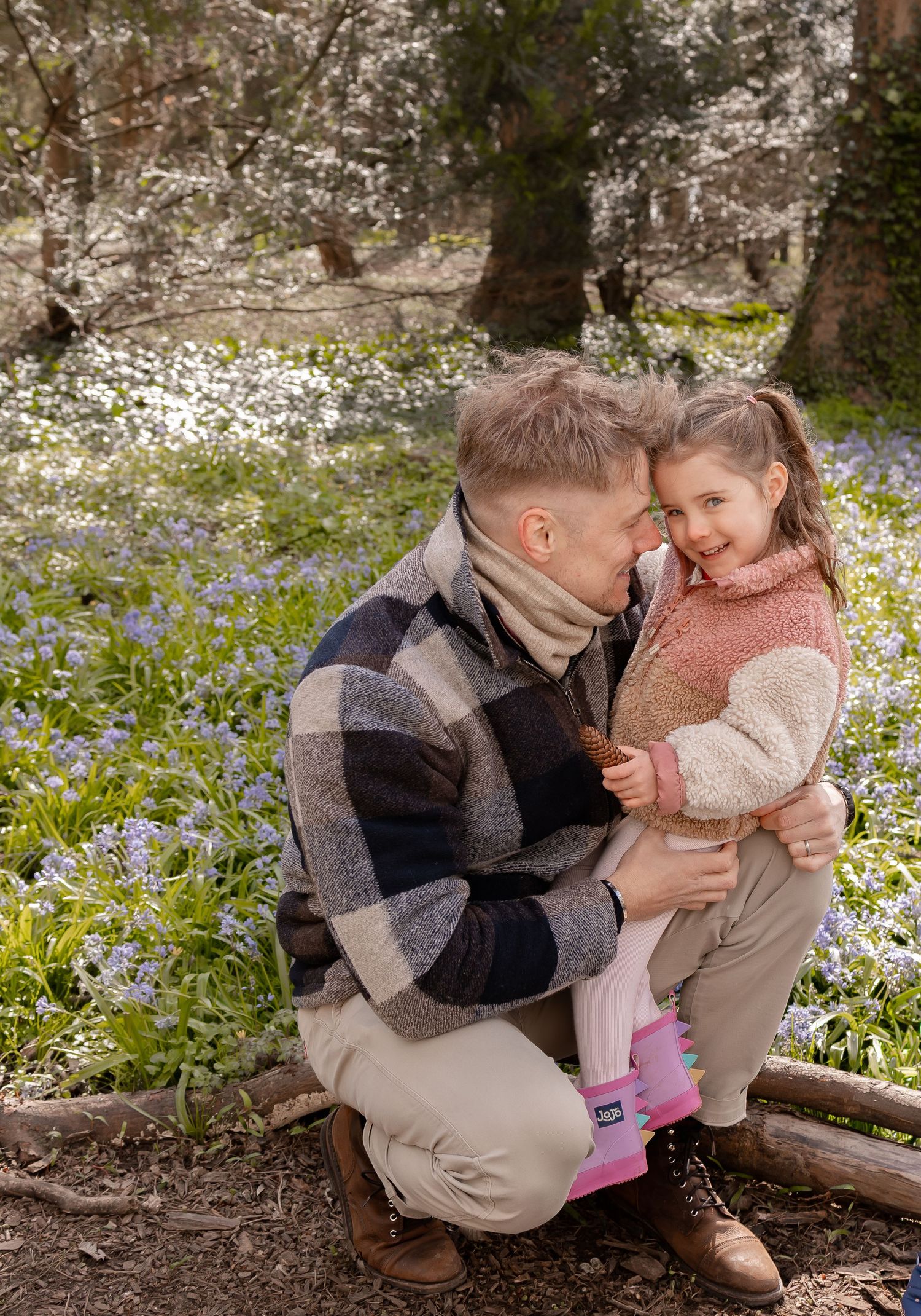 A father crouches to embrace his smiling young daughter among blooming wildflowers in a sunlit forest. Photography in Buckinghamshire.