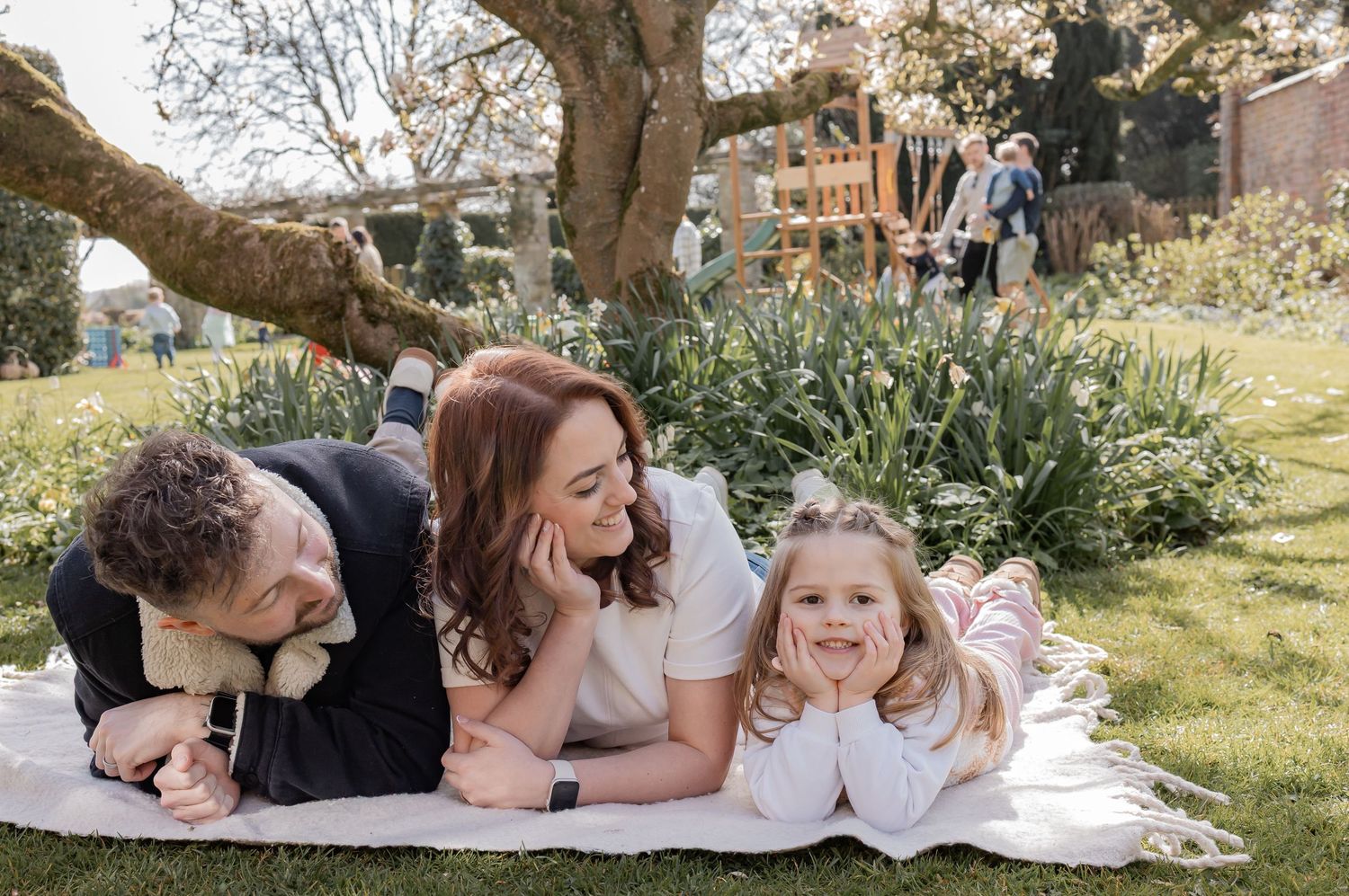 Family of three lying on a blanket in a garden, smiling together under a large tree on a sunny day. Photography in Buckinghamshire.