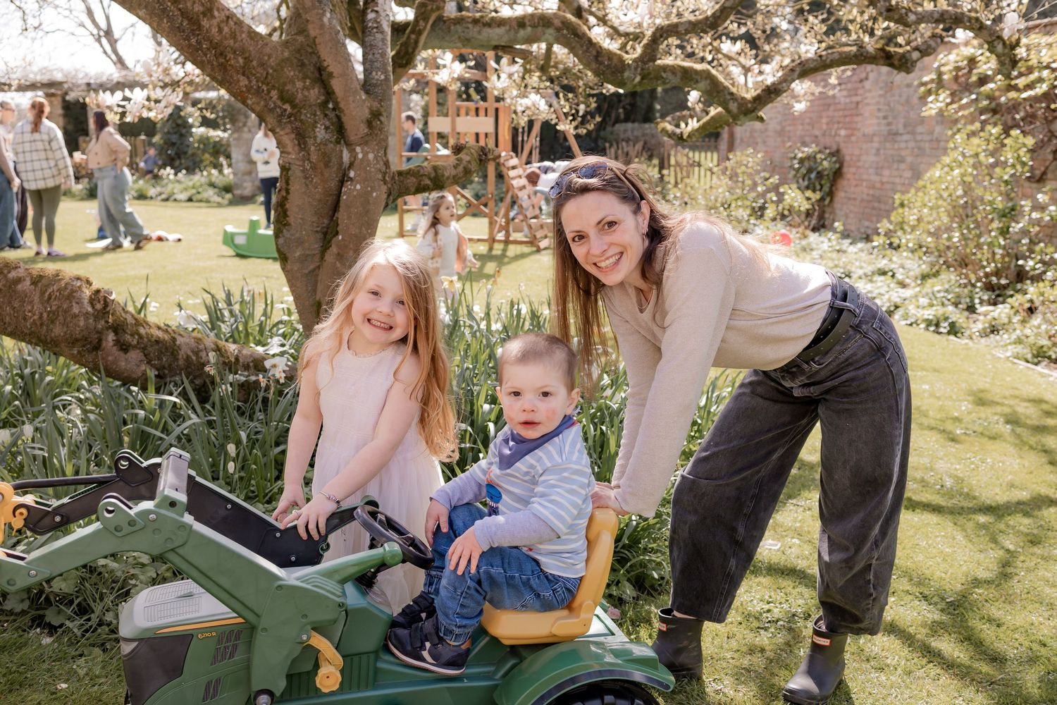 A woman and two young children pose with a green toy tractor in a sunny garden. Photography in Buckinghamshire.
