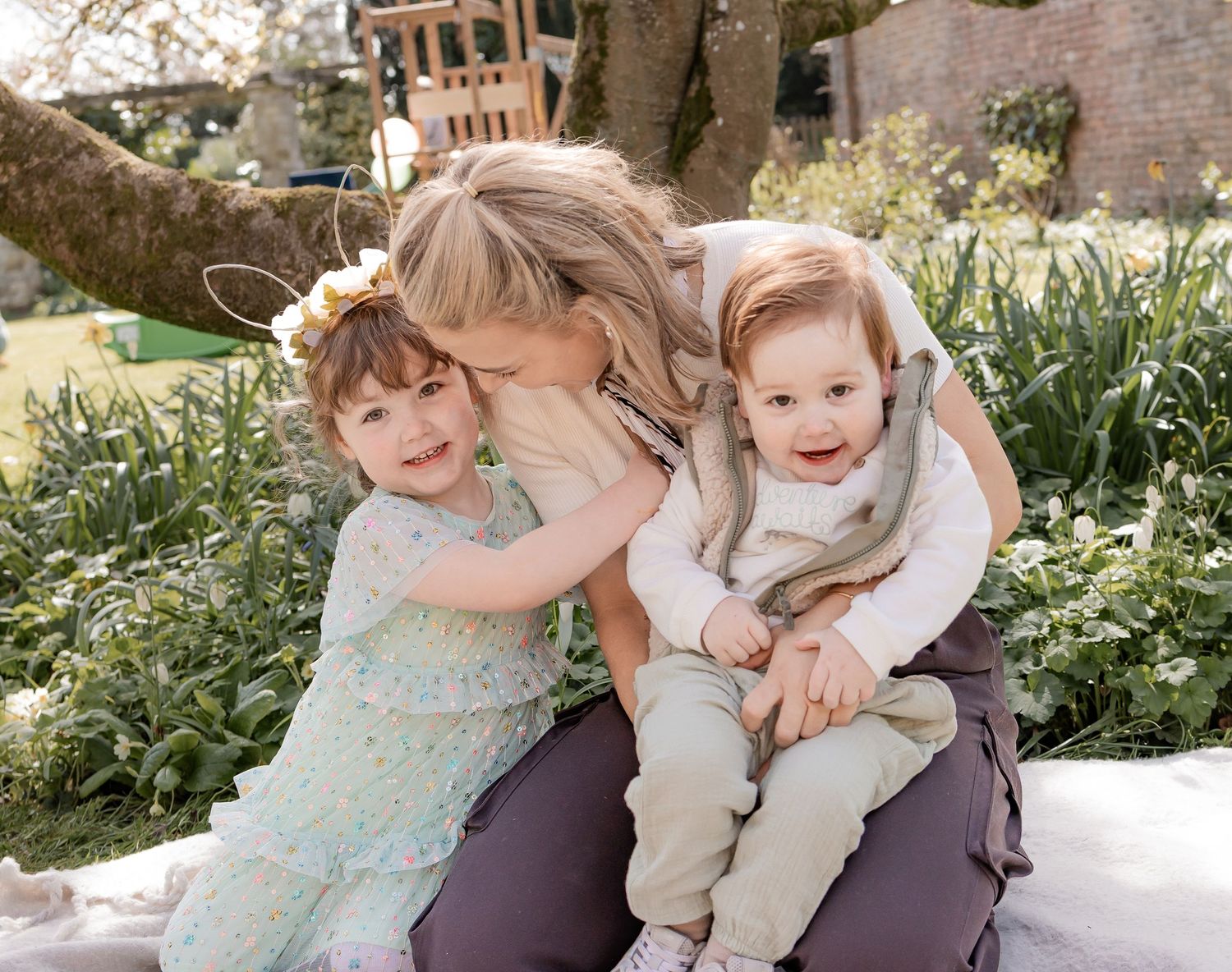 Blonde mother hugs and kisses two young children outdoors in a sunny spring garden setting. Photography in Buckinghamshire.