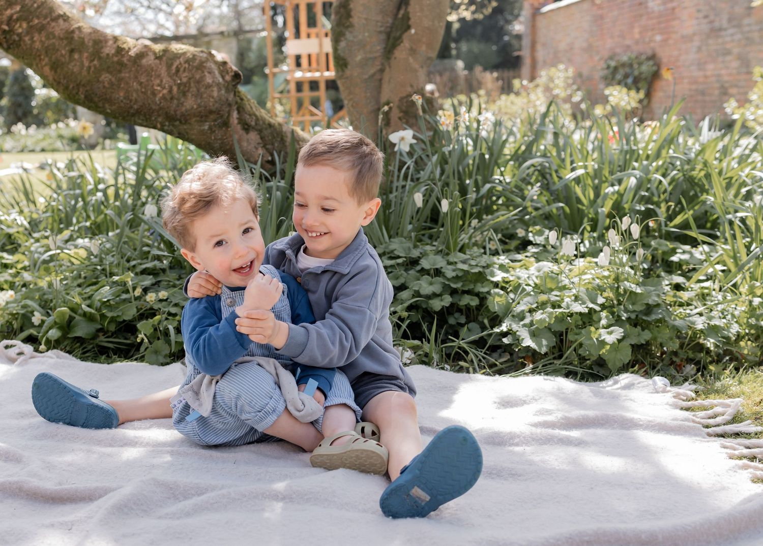 Two young brothers sit together laughing on a blanket in a beautiful spring garden. Photography in Buckinghamshire.
