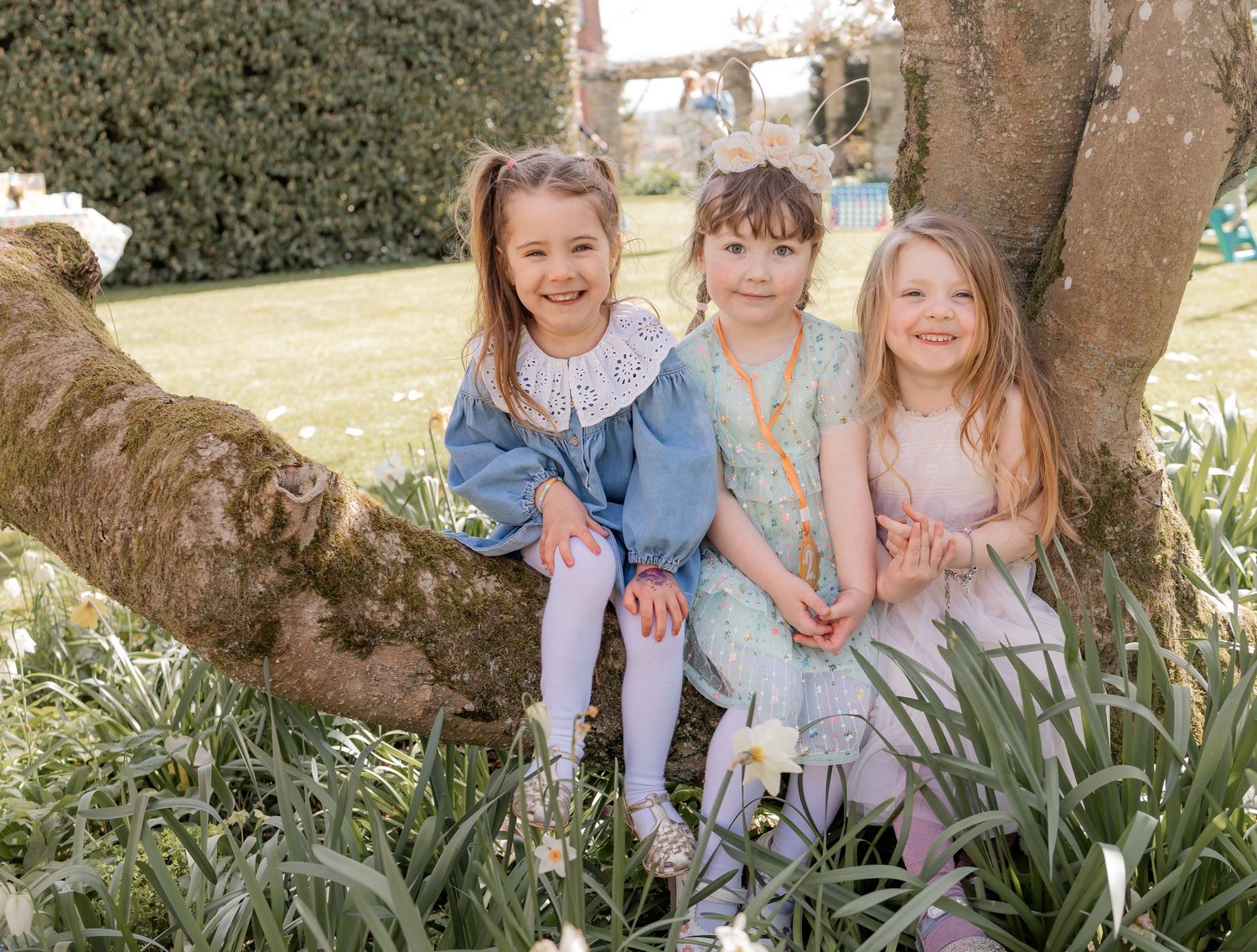 Three young girls smile and pose together by a tree trunk in a blooming spring garden. Photography in Buckinghamshire.