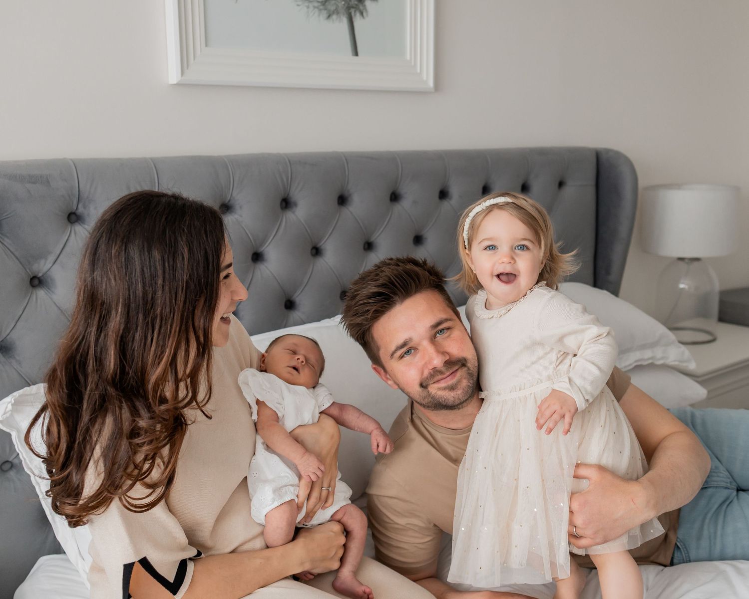 Happy family with newborn baby and toddler girl sitting on a grey tufted bed in a bright bedroom.