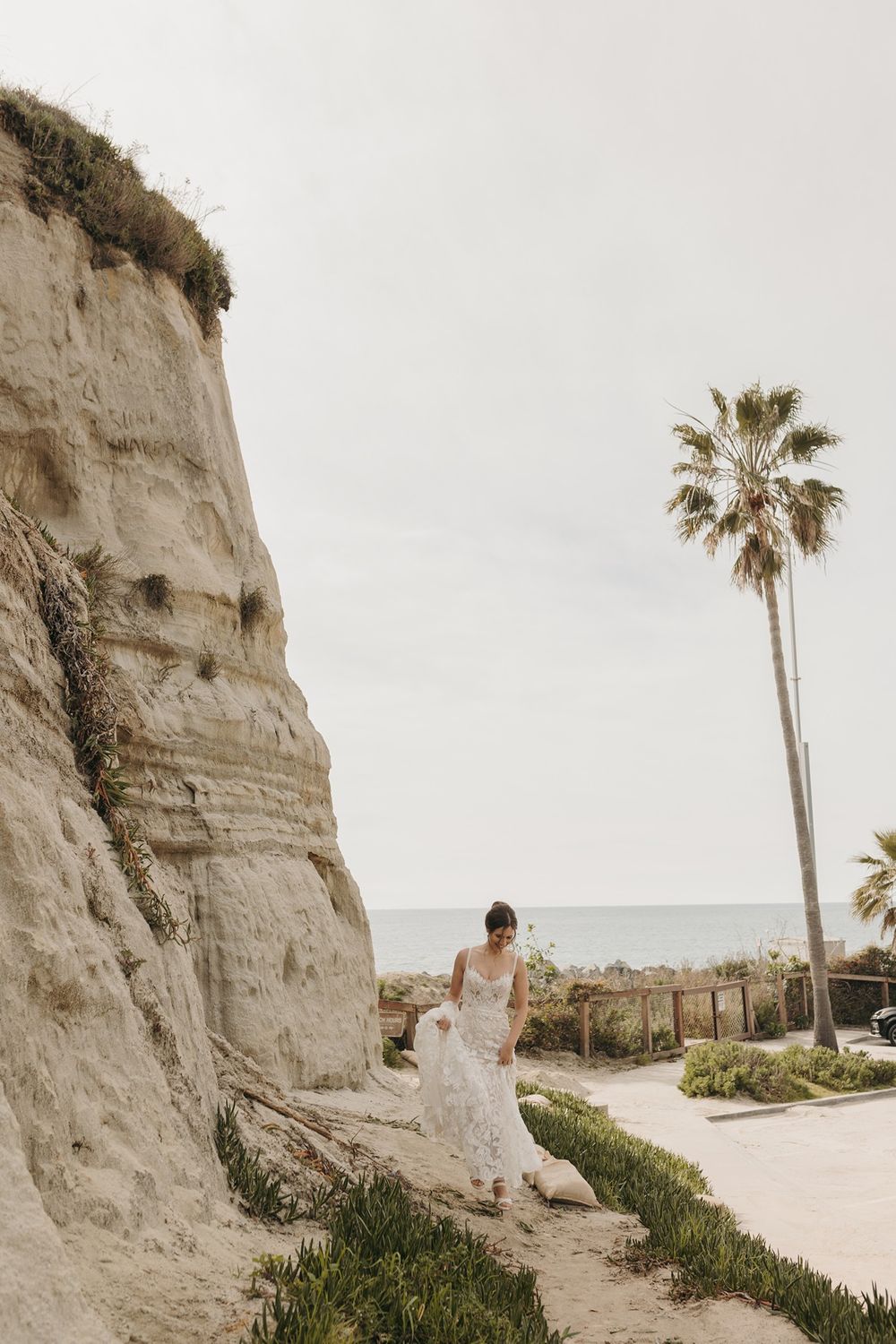 A bride walks down a beachside path in a dreamy coastal scene against limestone cliffs and palm trees at Calafia Beach Park in San Clemente.
