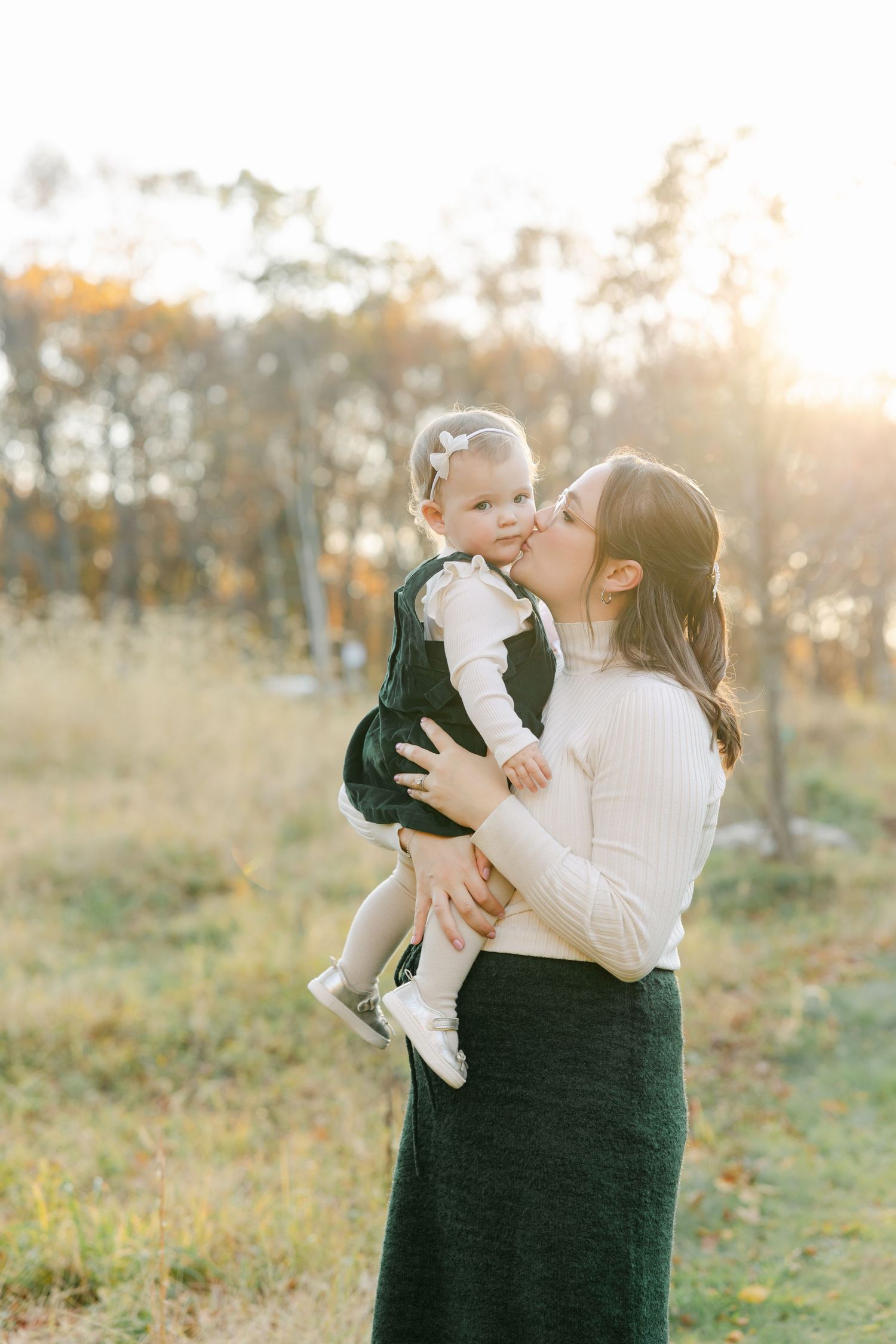 Mother kissing baby girl outdoors in golden sunset light, both dressed in dark and white outfits in autumn park.