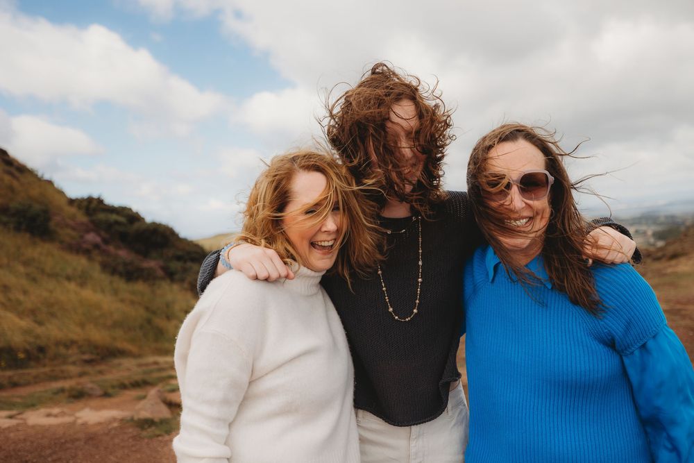 Three friends smile together on a hiking trail with rolling hills and cloudy skies in the background.