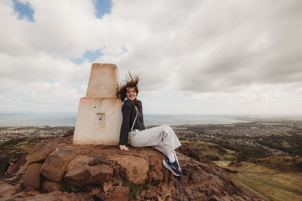 A person sits on a rocky outcrop overlooking a vast desert landscape under cloudy skies.
