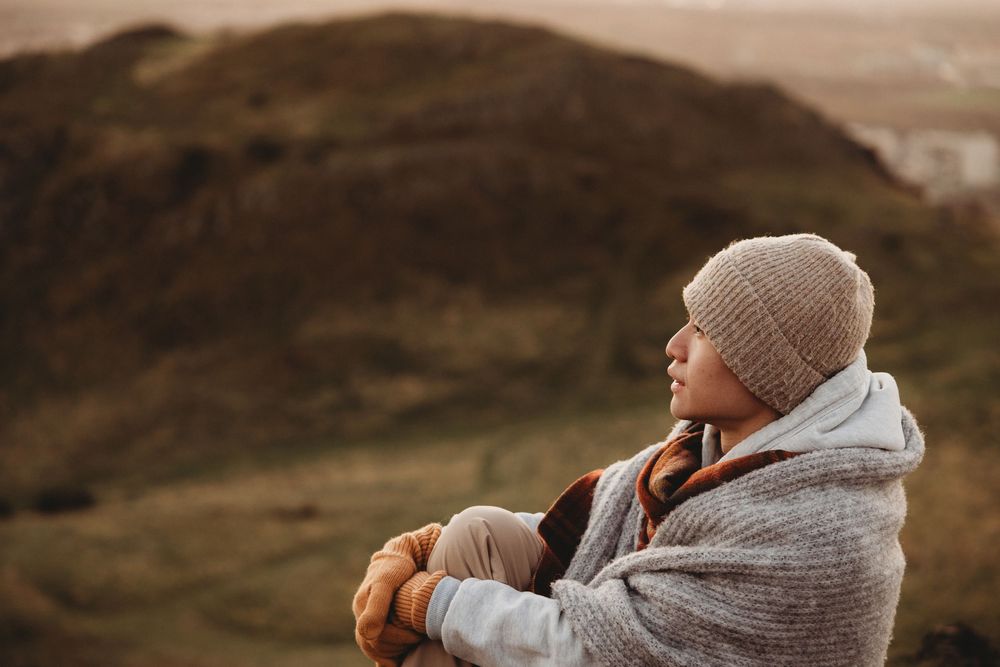 A couple in warm winter clothing sits together on a hillside overlooking a scenic mountain landscape at sunset.