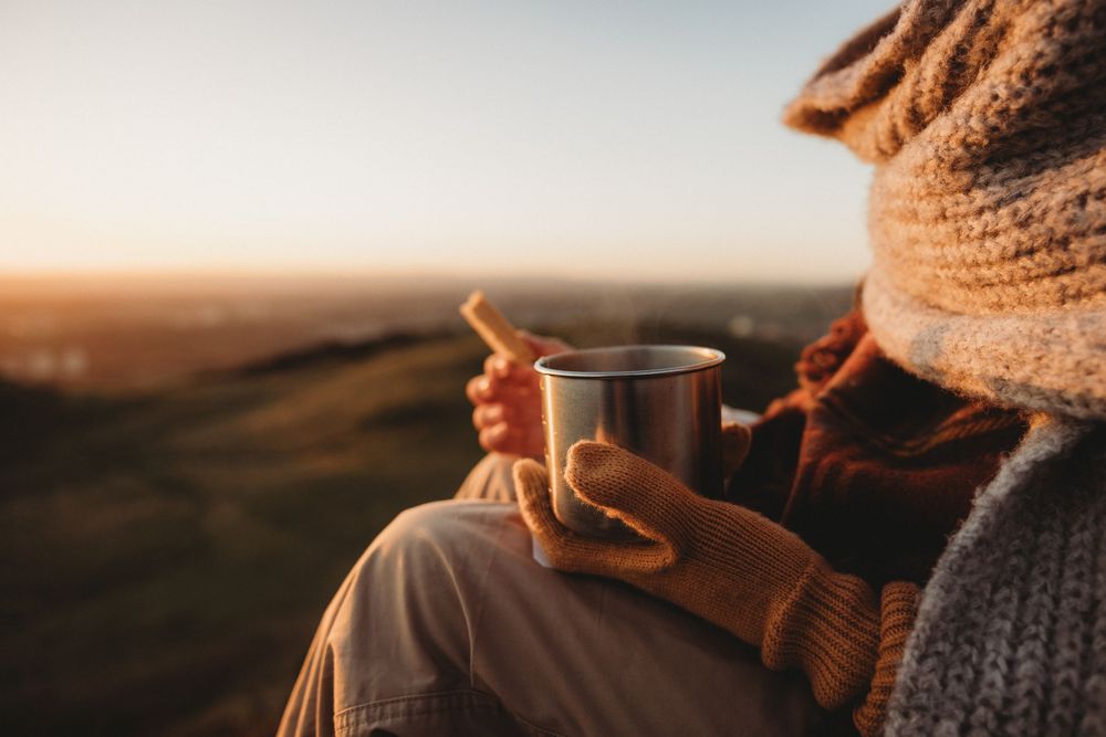 Person wearing cozy knit sweater holds steaming coffee mug while enjoying sunset view outdoors.