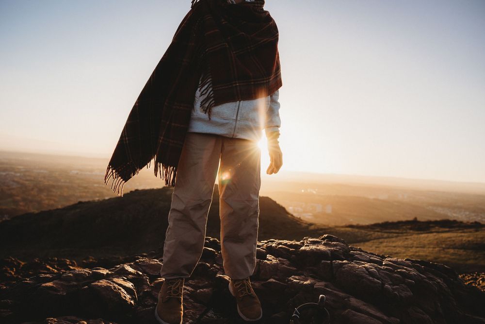 Hiker stands on rocky mountain peak at sunset with jacket and backpack overlooking vast wilderness landscape.