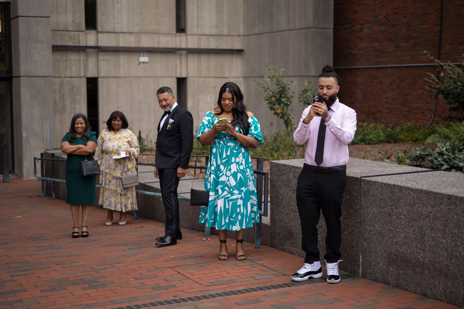 Panoramic view of people sitting and standing along a brick wall outside a building entrance.