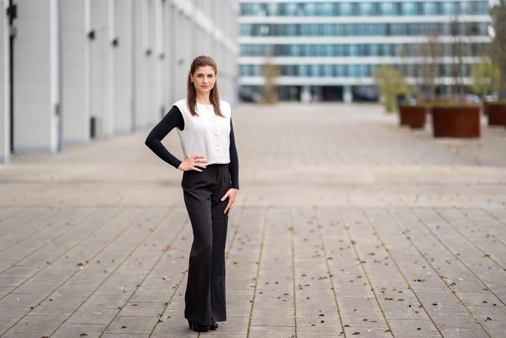 Professional woman in business attire stands confidently with hand on hip in modern office setting.