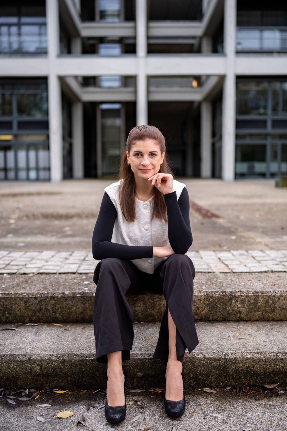 Professional in business attire sits on concrete steps outside modern office building wearing black and white outfit.