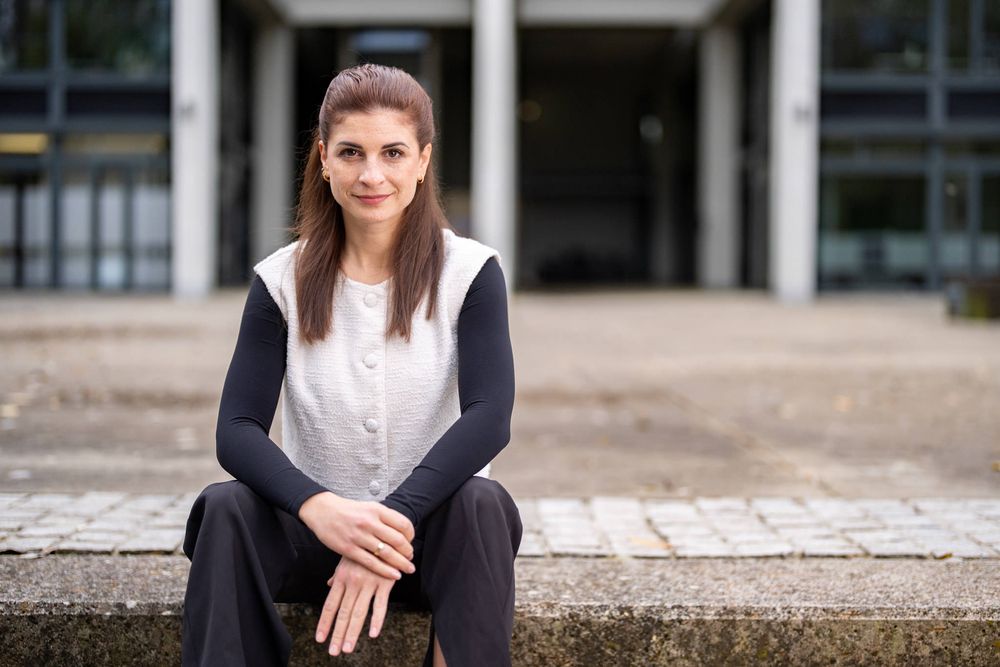 Woman in navy blazer and white shirt seated outdoors with a confident expression against an urban setting.