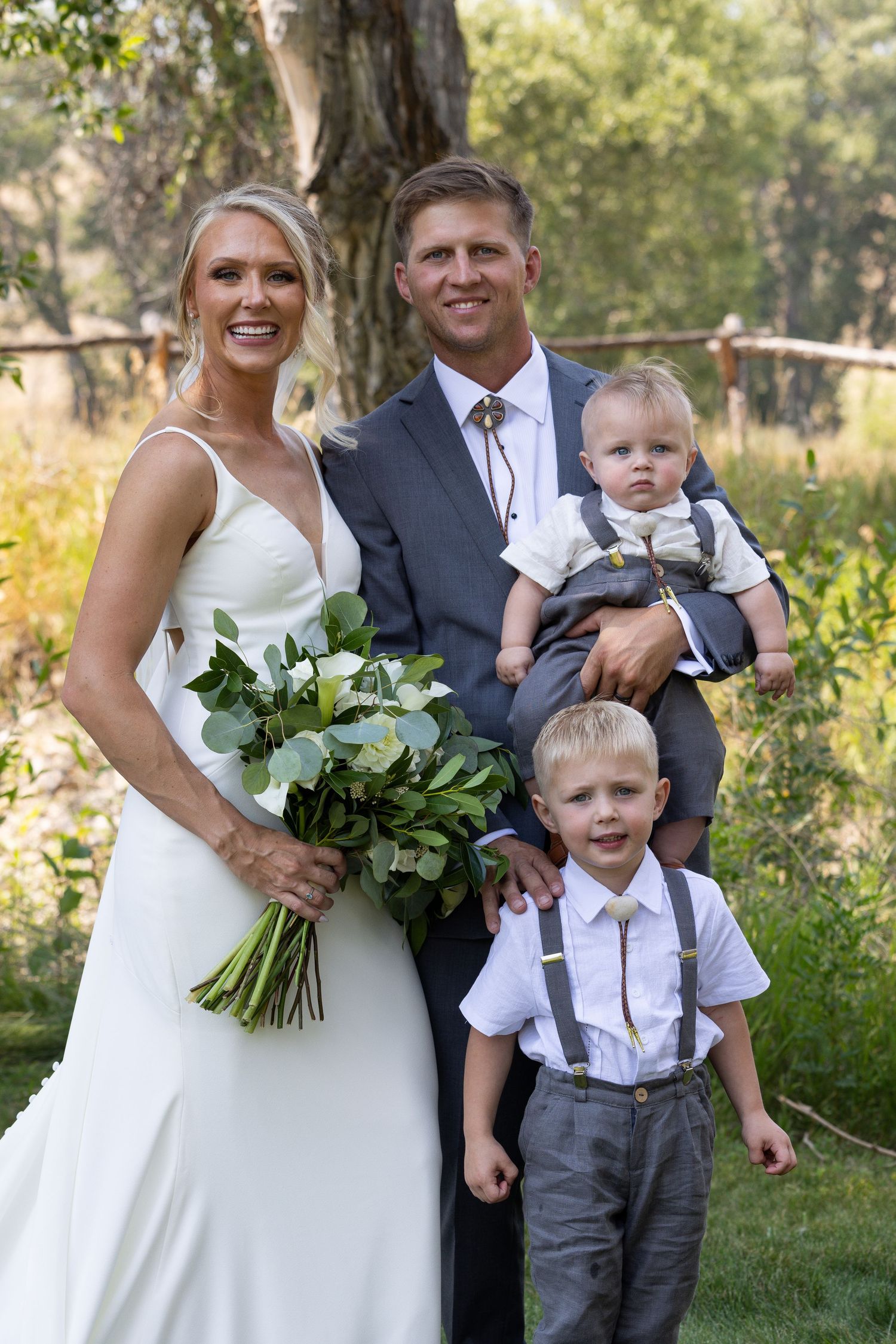 Wedding party poses outdoors in formal attire with white bouquet and grey suits in natural garden setting.