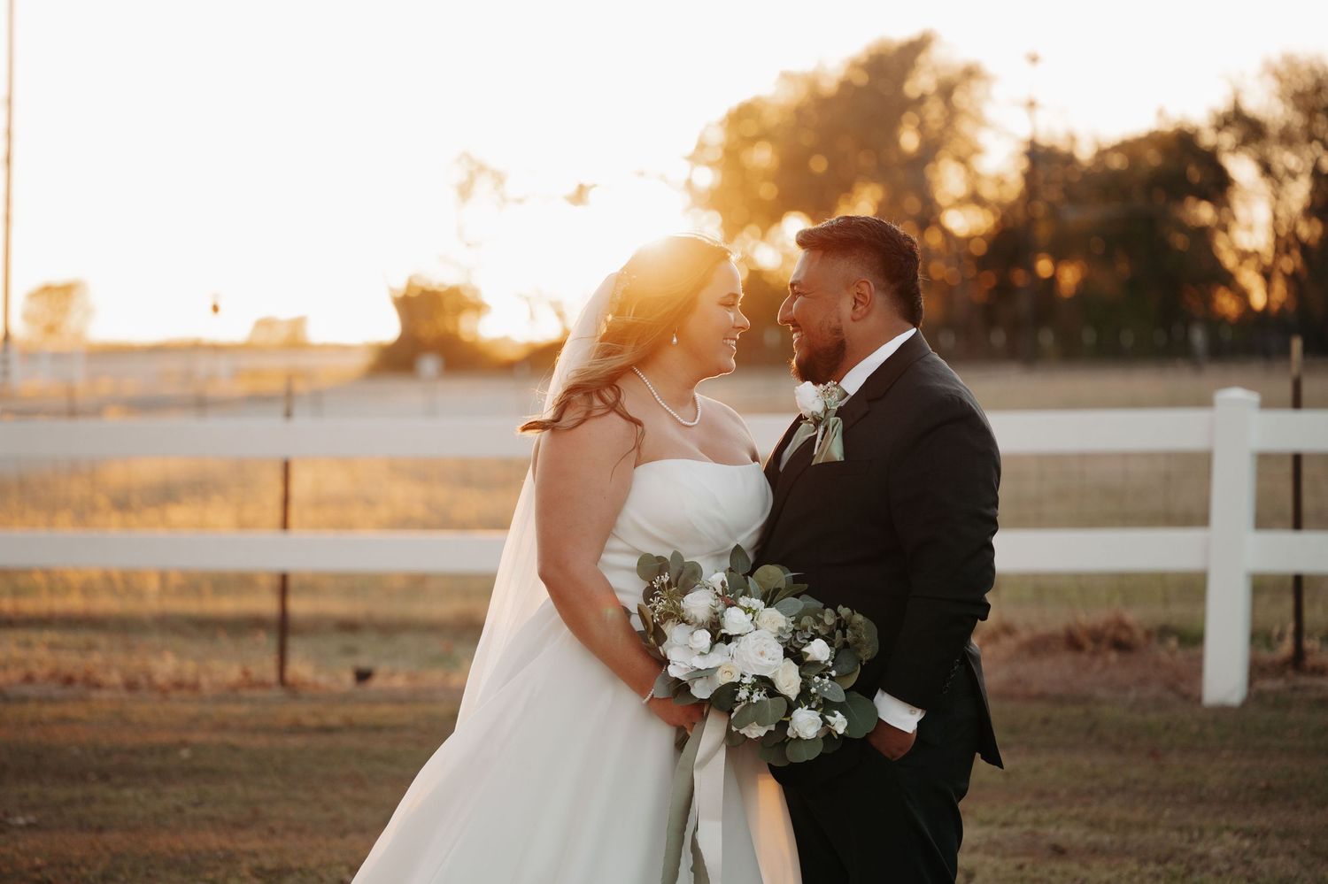 Romantic wedding couple embraces during golden hour sunset by a white fence at a rustic outdoor venue.
