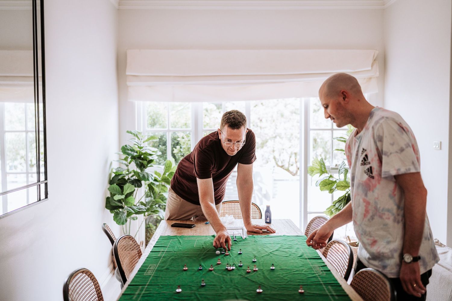 A group of people playing foosball on a green table in a bright, sunlit room.