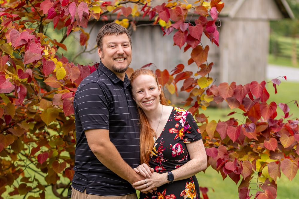 Two people embrace and smile while standing in front of vibrant red and orange autumn foliage.