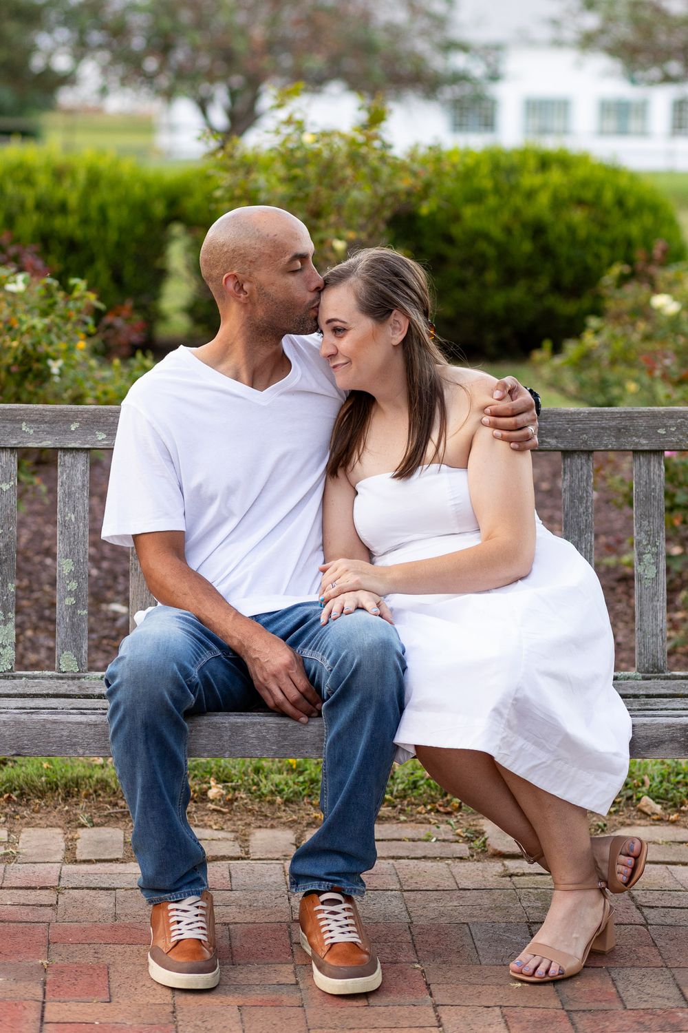 A couple shares an intimate moment while sitting on a wooden bench in a garden with blooming flowers and greenery.