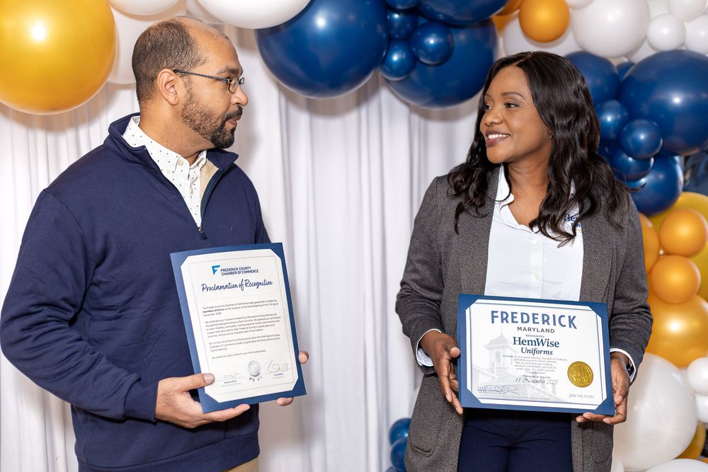 Two people holding award certificates smile at each other in front of a blue and gold balloon backdrop.