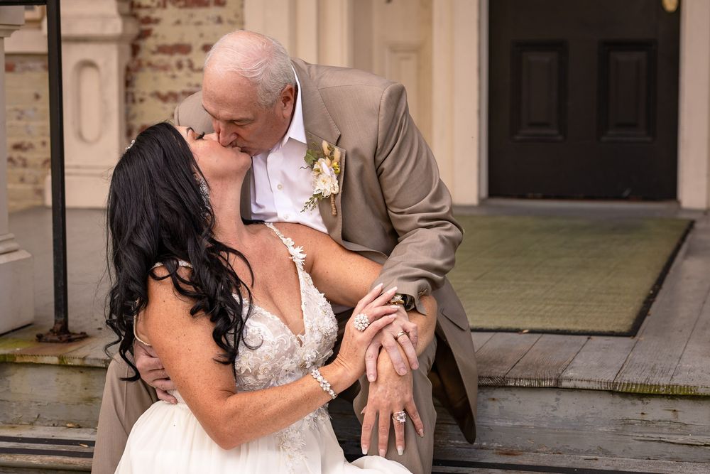 A couple shares a romantic kiss in wedding attire on the steps of a historic mansion.