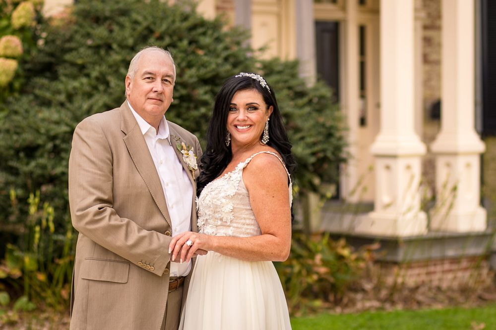 A couple poses together in wedding attire outside a historic building with stone columns and greenery.
