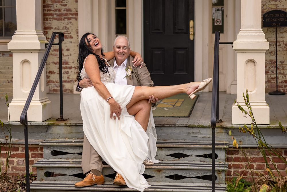 A wedding couple shares a playful moment on the steps of a brick building with white columns and a black door.