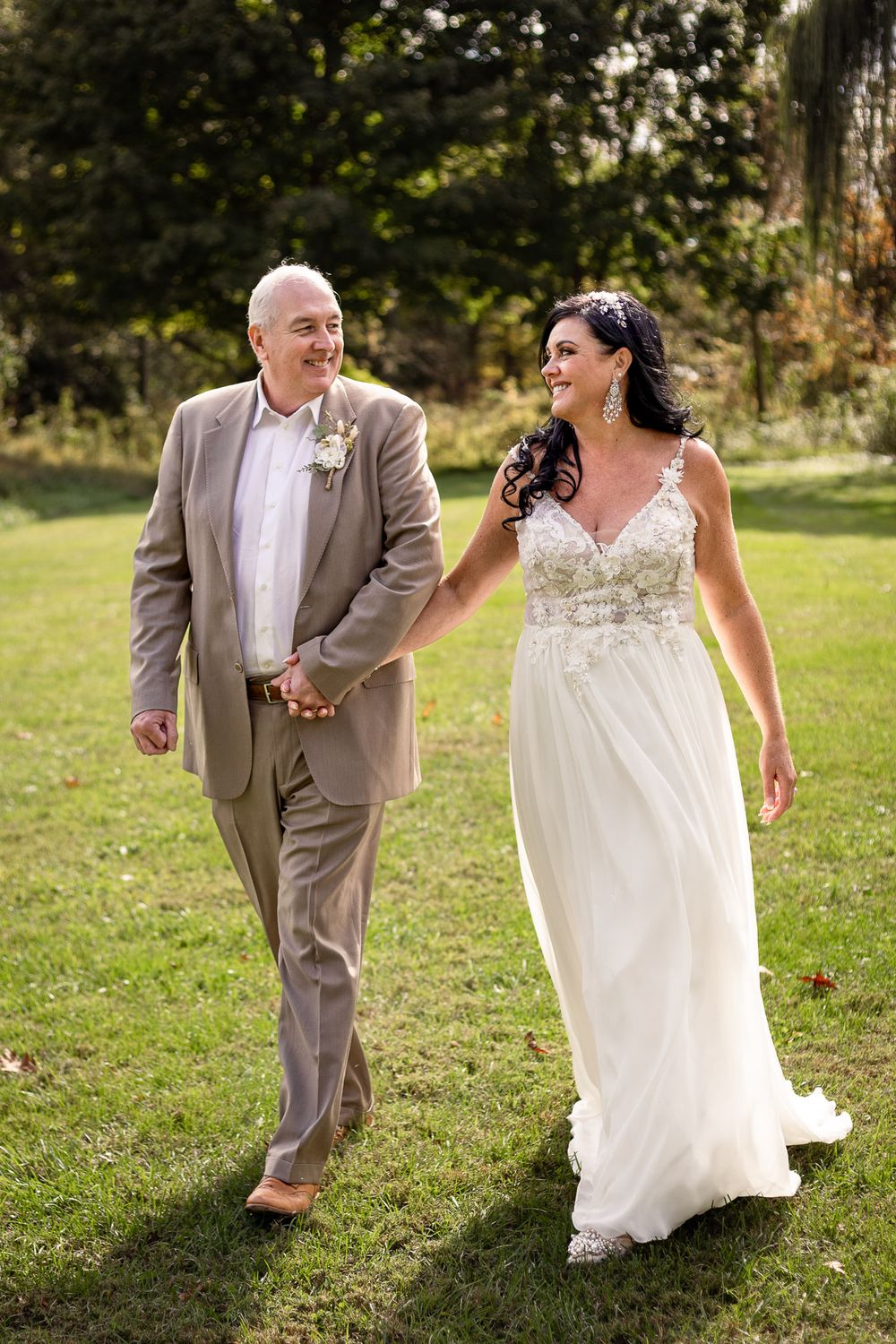 A couple in wedding attire walks and laughs together on a grassy lawn during their outdoor wedding celebration.