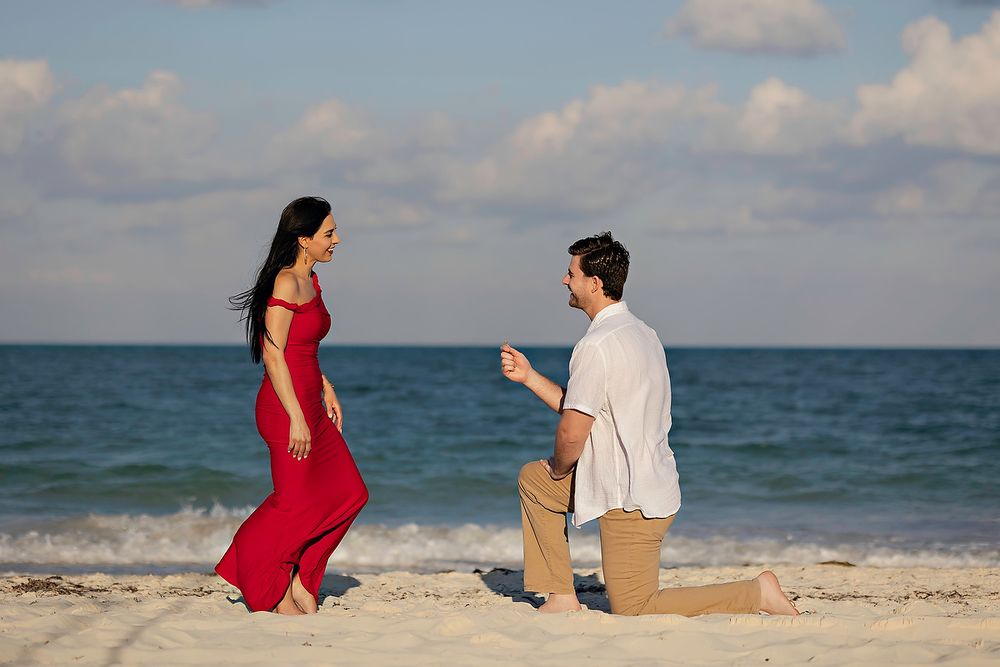 A romantic beach proposal scene with a person in a red dress and another kneeling on the sand at sunset.