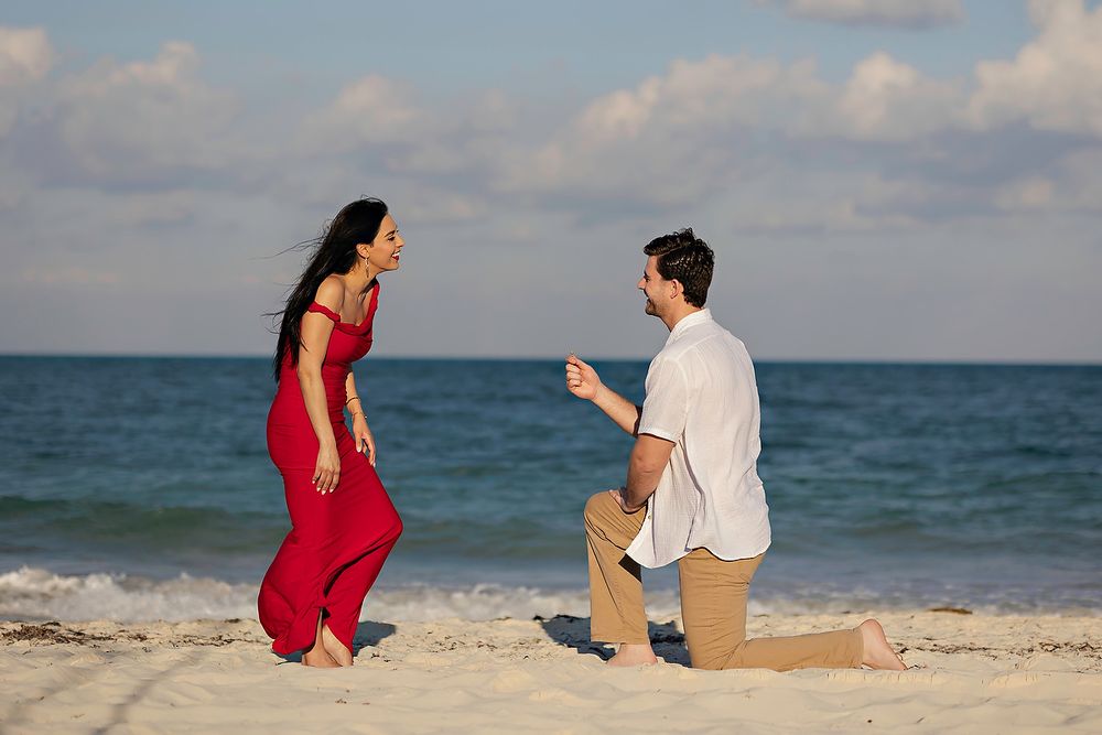 A couple sits together on a sandy beach with the blue ocean and cloudy sky in the background.