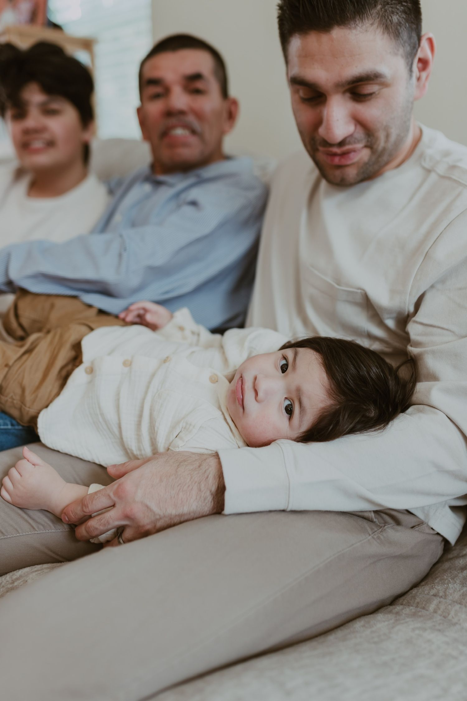 Family relaxing on couch with baby lying across laps, looking up at camera in cozy home setting.