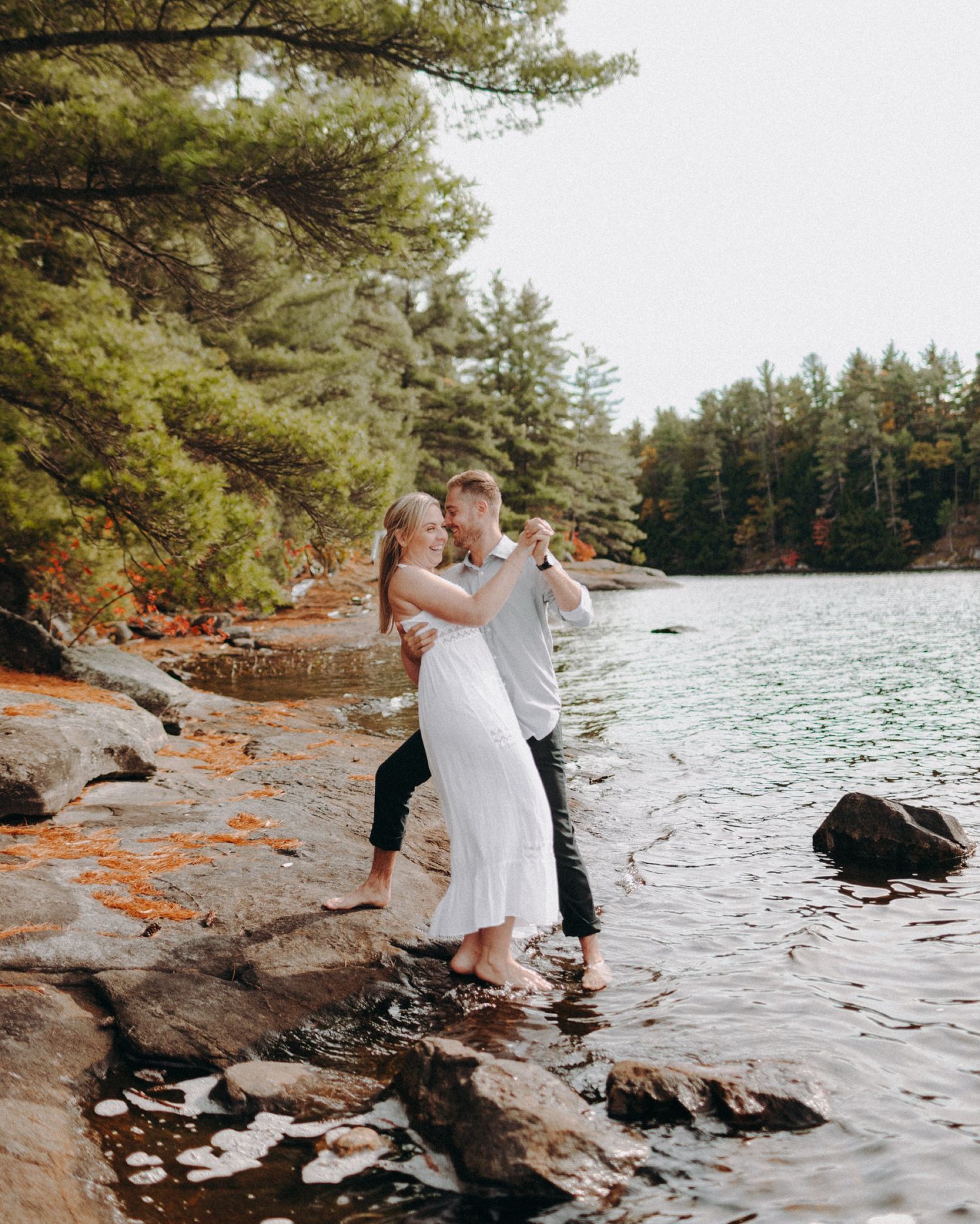 A couple in wedding attire dances together on a rocky shoreline beside a serene lake surrounded by pine trees.