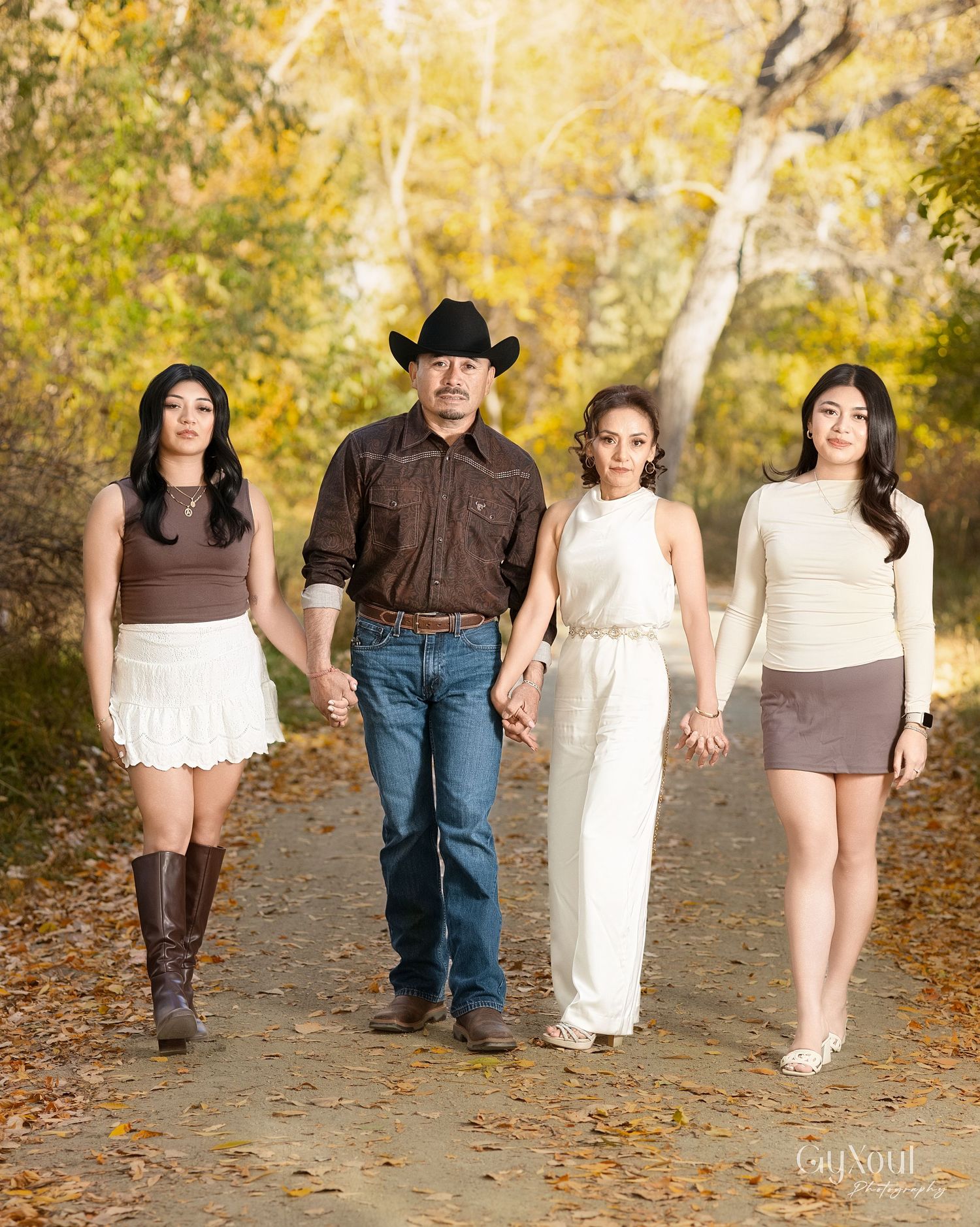A family walks together on a leaf-covered autumn path through golden trees wearing coordinated brown and white outfits.