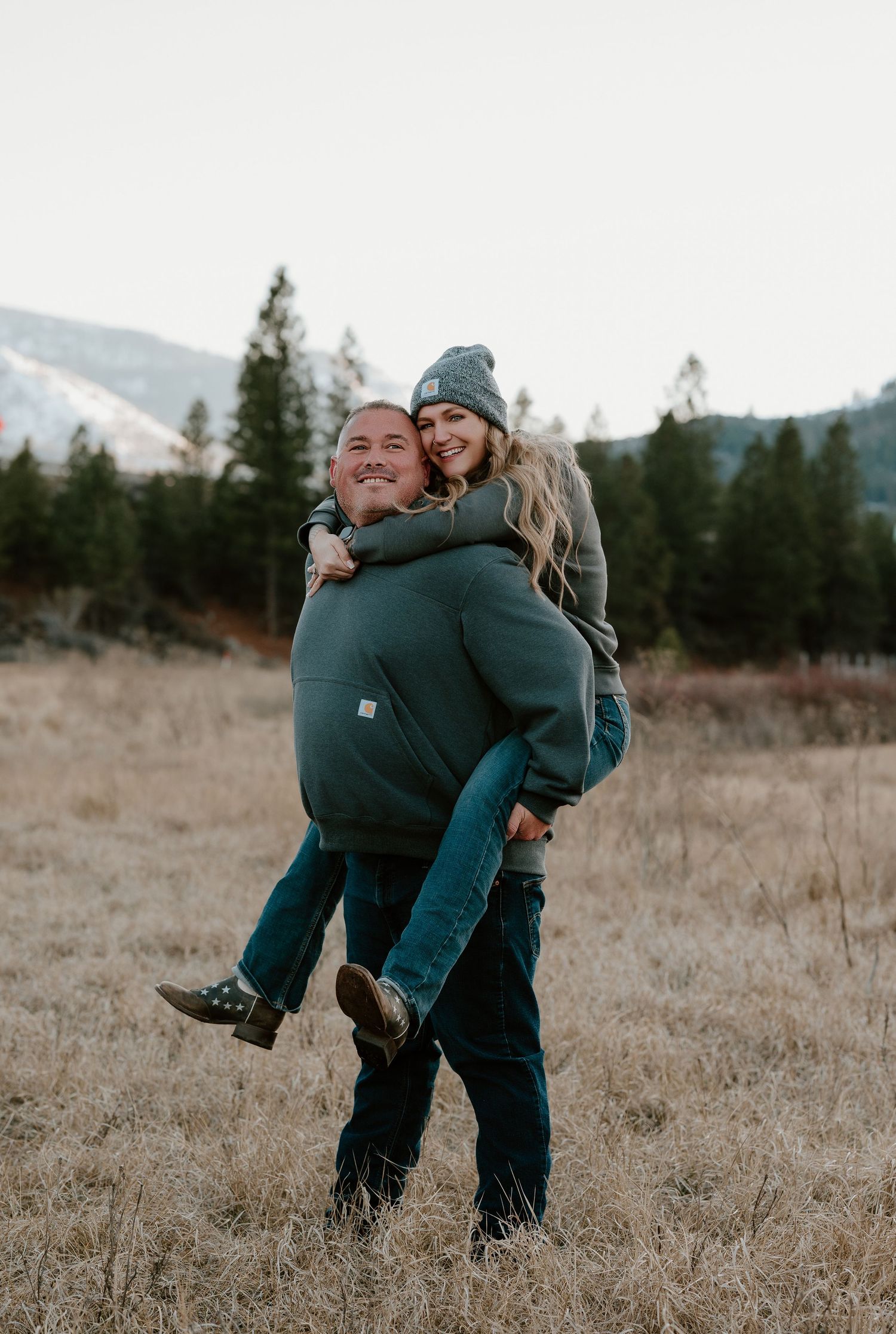 Couple playfully embracing and spinning in a golden field with mountains and evergreen trees in the background.