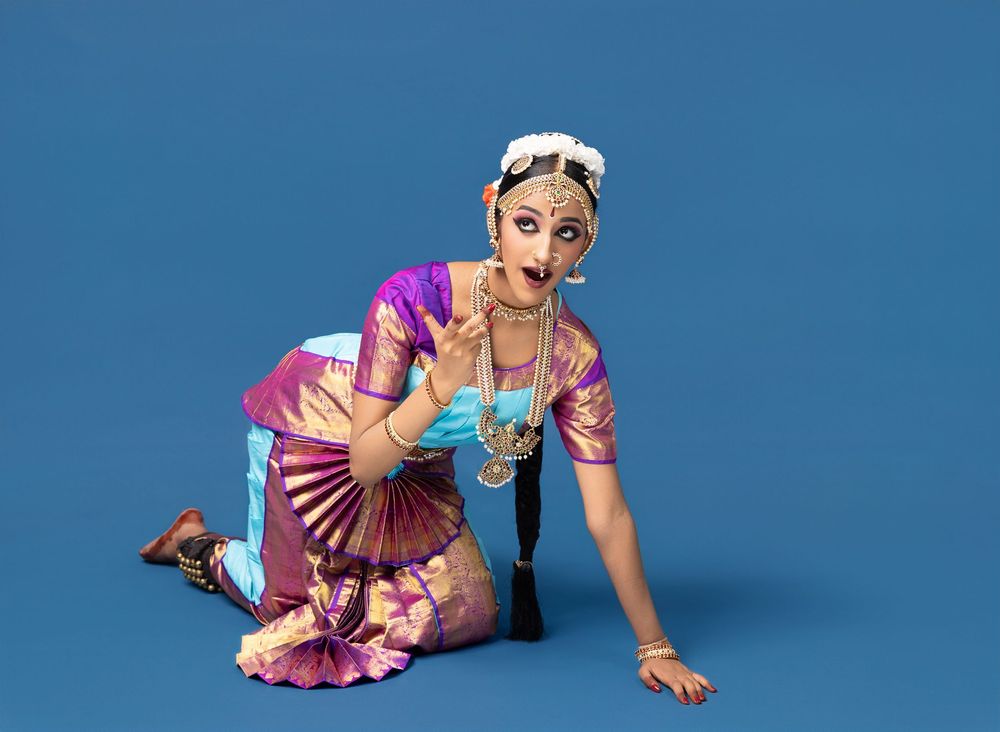 Dancer in vibrant pink and purple traditional Indian dance costume poses gracefully against blue background.
