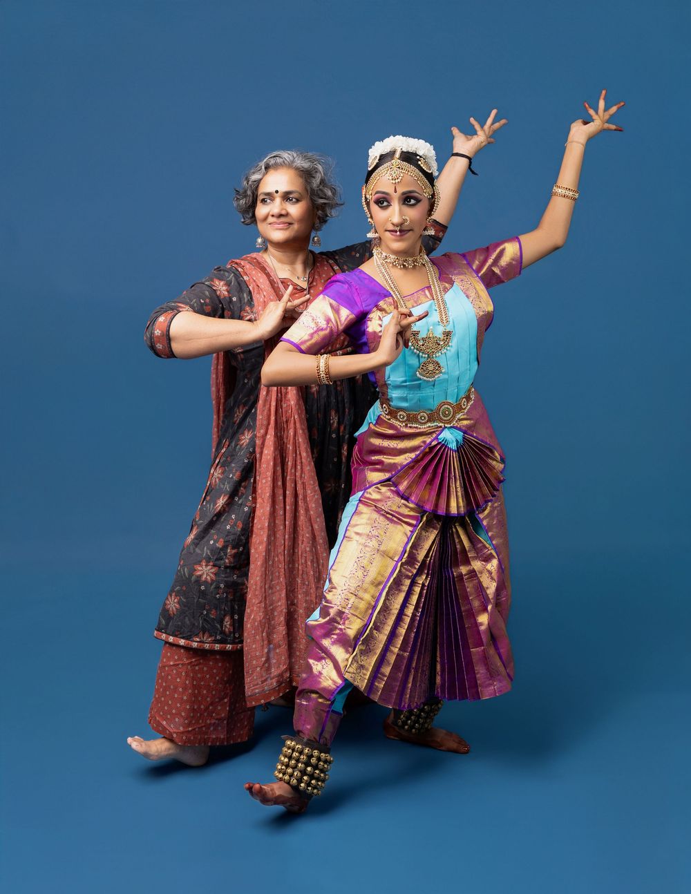 Two dancers in traditional Indian attire perform graceful movements against a blue backdrop in a series of poses.
