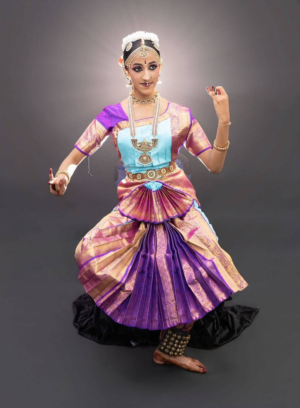A dancer in traditional Indian dance attire with colorful silk costume and jewelry performs classical dance poses against gray backdrop.
