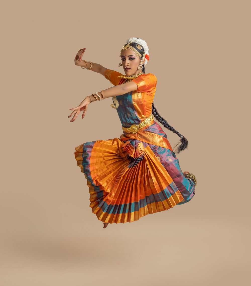 Dancer in vibrant orange and blue traditional Indian dance costume performs graceful Bharatanatyam movements.