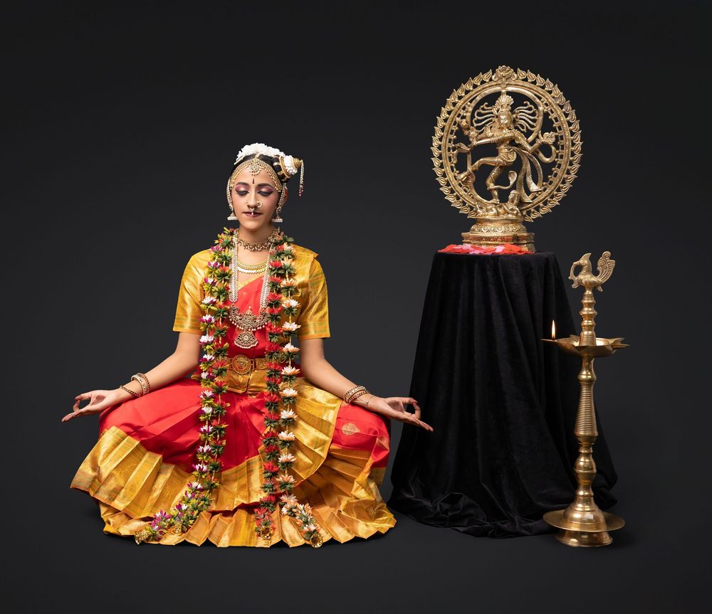 Traditional Indian classical dancer in vibrant yellow and red costume performs with traditional brass lamp on dark stage.
