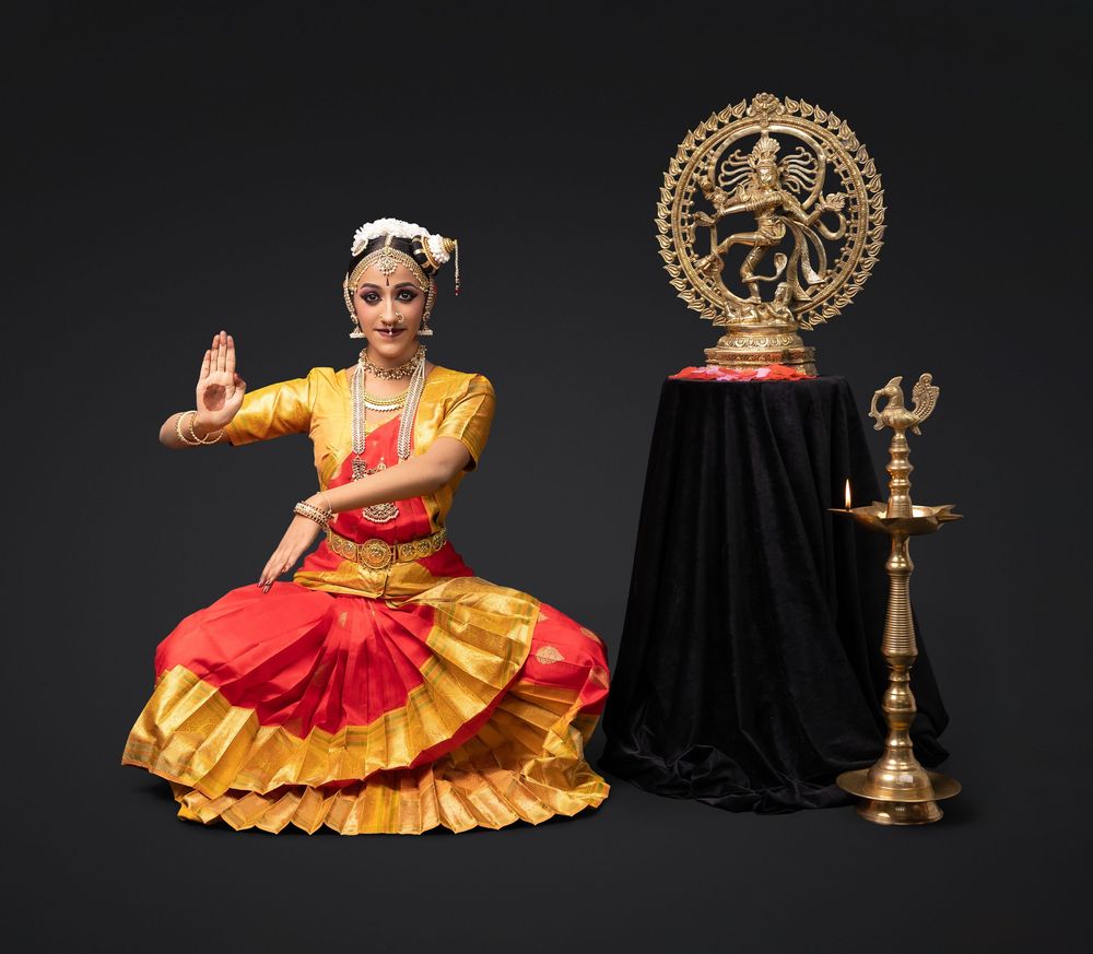Classical Indian dancer in red and gold costume performs traditional Bharatanatyam dance poses with traditional oil lamp.