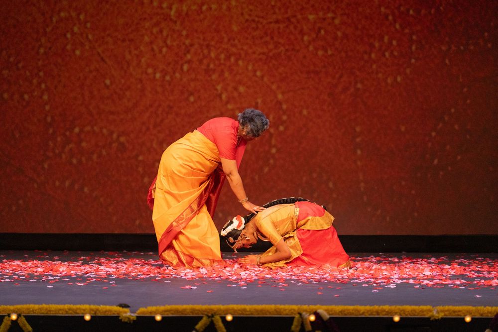 A dancer in orange costume performs a classical Indian dance sequence on a stage with red lighting effects.