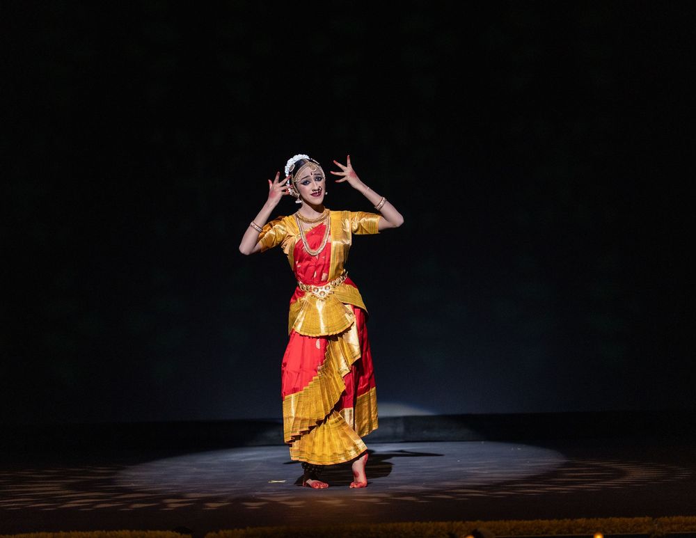 A performer in traditional yellow Indian dance attire performs expressive movements on a dimly lit stage.