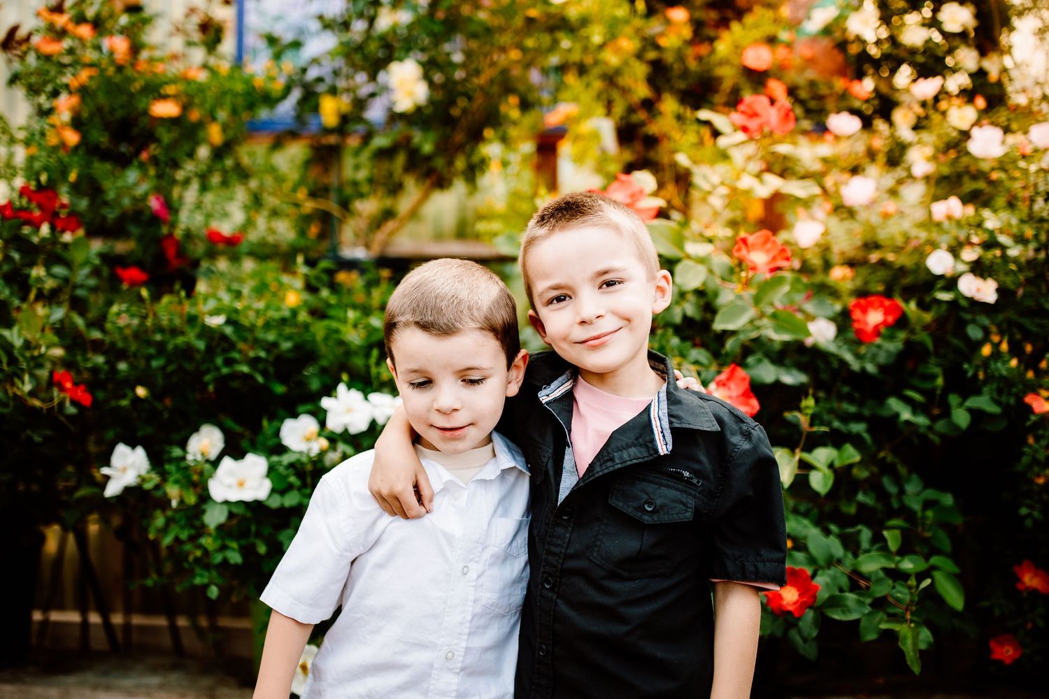 Two friends stand together in front of colorful floral garden on sunny day.