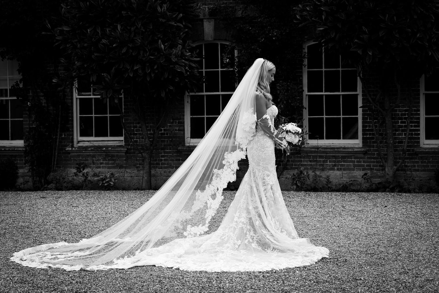 A bride in an elegant lace wedding gown with a dramatic long veil poses on a lawn in front of a syrencot, cotswolds wedding venue